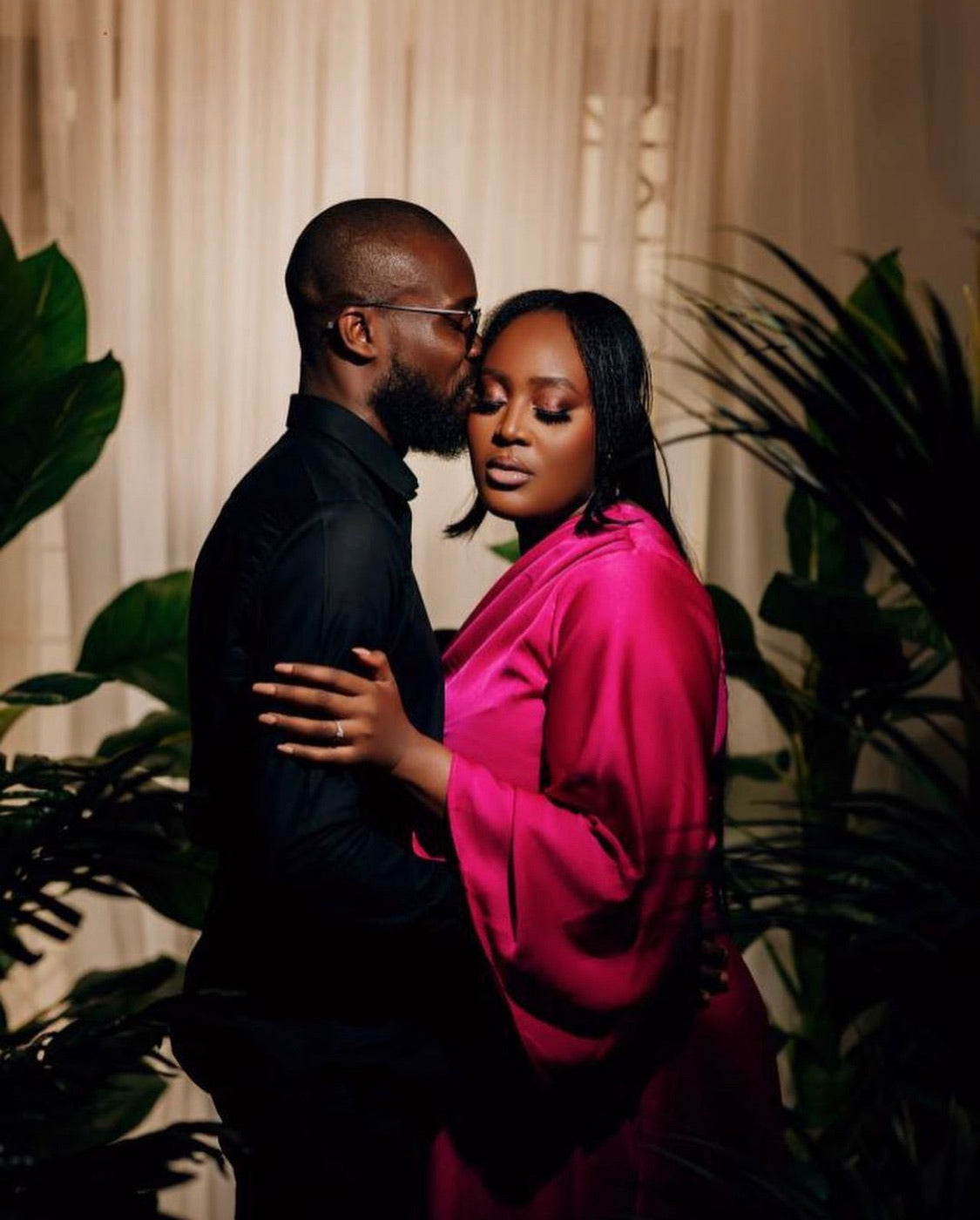 Couple embracing, man in black shirt and woman in pink satin dress indoors with plants