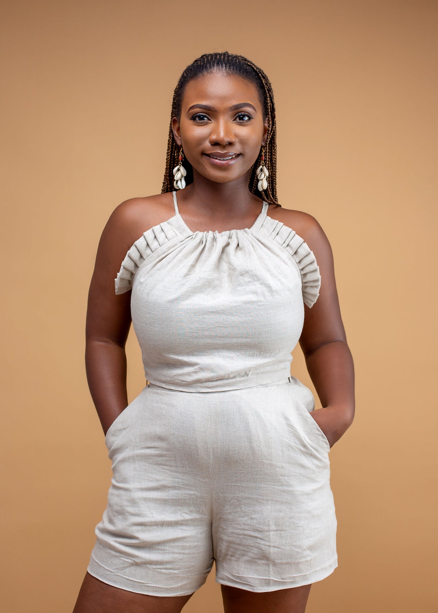 Smiling woman in beige sleeveless romper with side pockets and shell earrings, neutral background