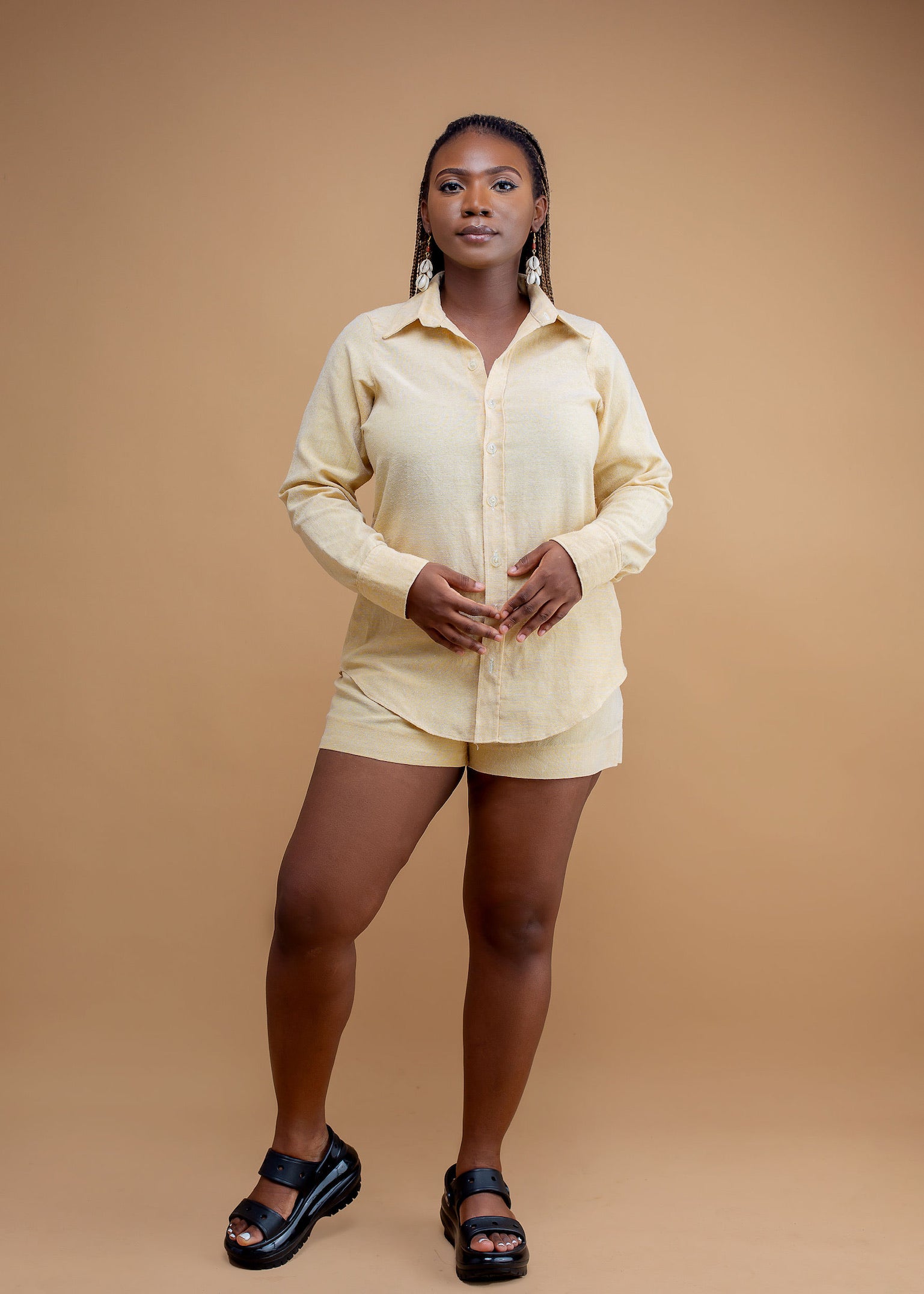 Woman wearing a light yellow button-up shirt and shorts set with black sandals, studio background