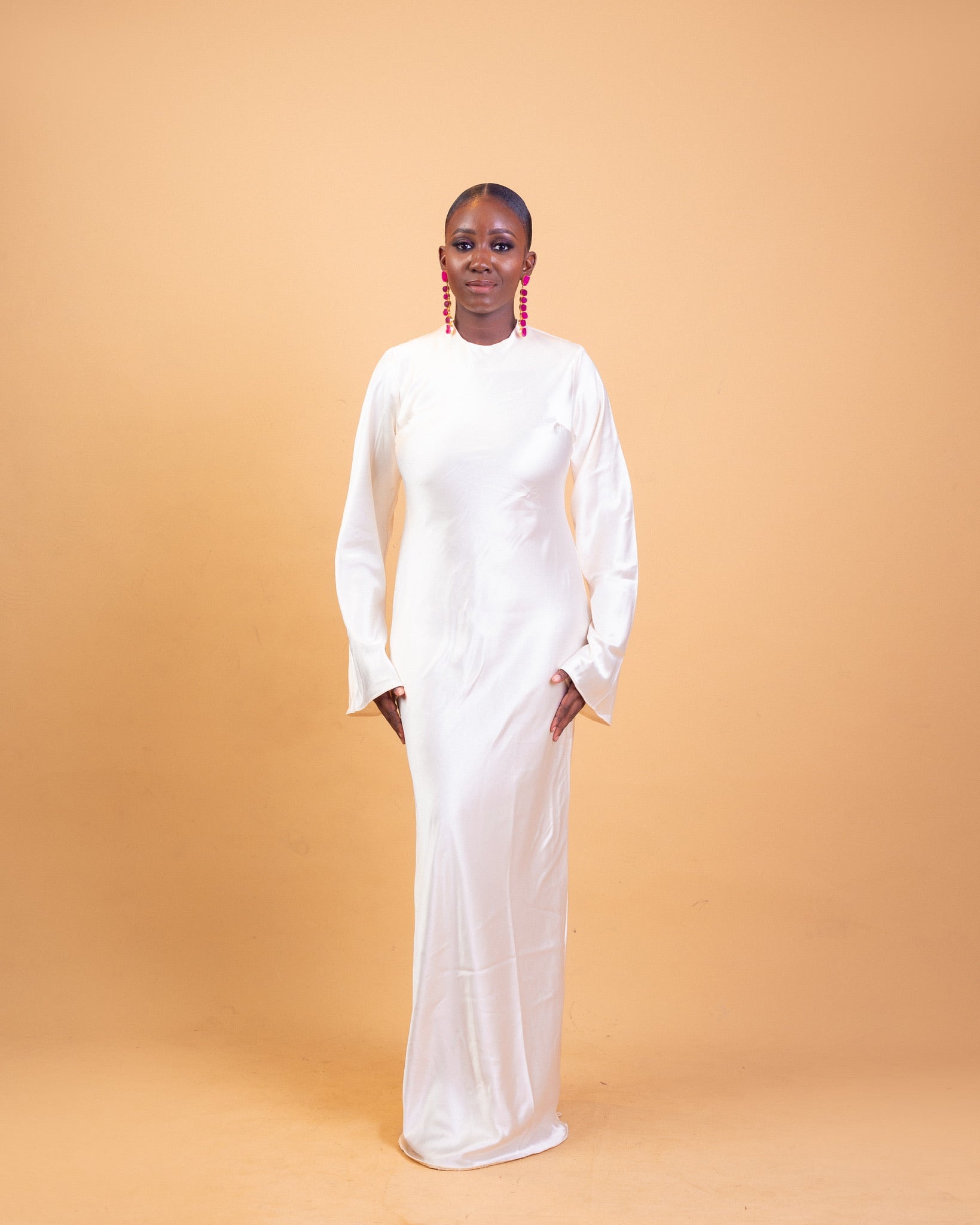 Woman in a long white gown with red earrings, posing against a peach background