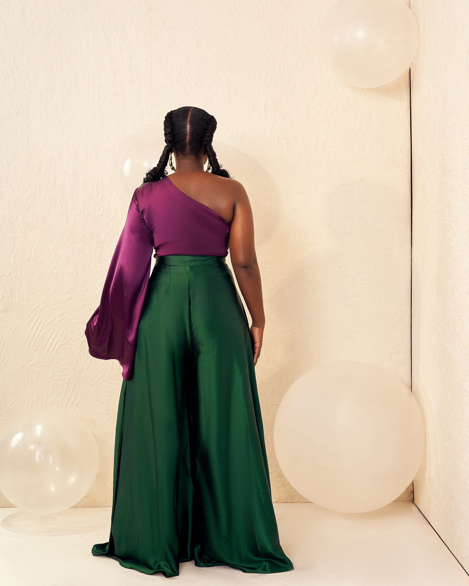 Woman in a purple one-shoulder top and green wide-leg pants, standing with balloons